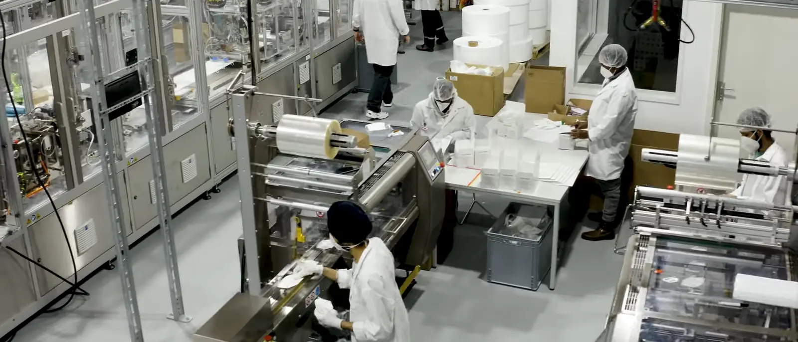 Fertigungslinie von Your Mask: Mitarbeitende in Schutzkleidung an Maschinen zur Herstellung und Verpackung von FFP2-Masken, im Hintergrund Rollen mit Vliesstoff