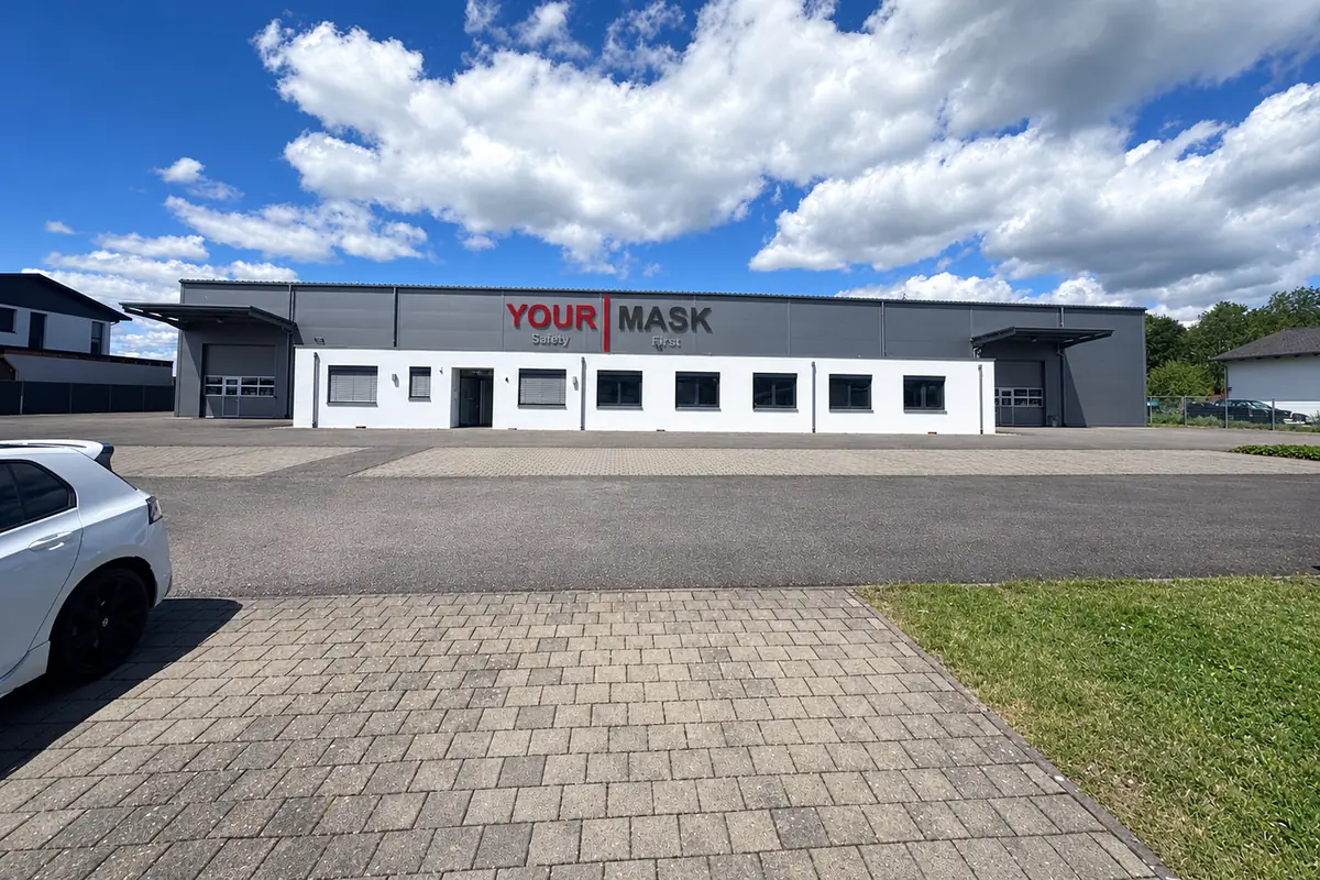 KI-generierte Ansicht des Produktionsstandorts von Your Mask in Bad Dürrheim: Fertigungshalle mit Büroanbau und dem Schriftzug YOUR MASK Safety First an der Fassade
